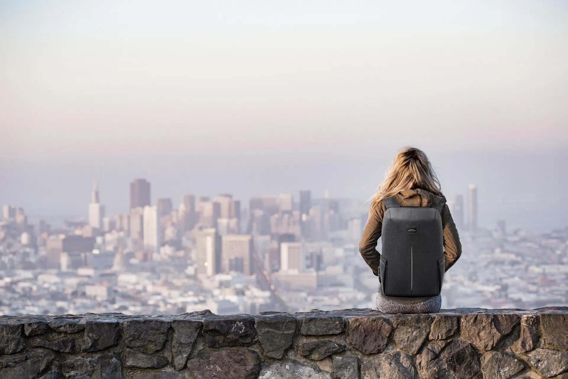 Frau mit Rucksack sitzt auf einer Mauer mit Blick auf die Stadt
